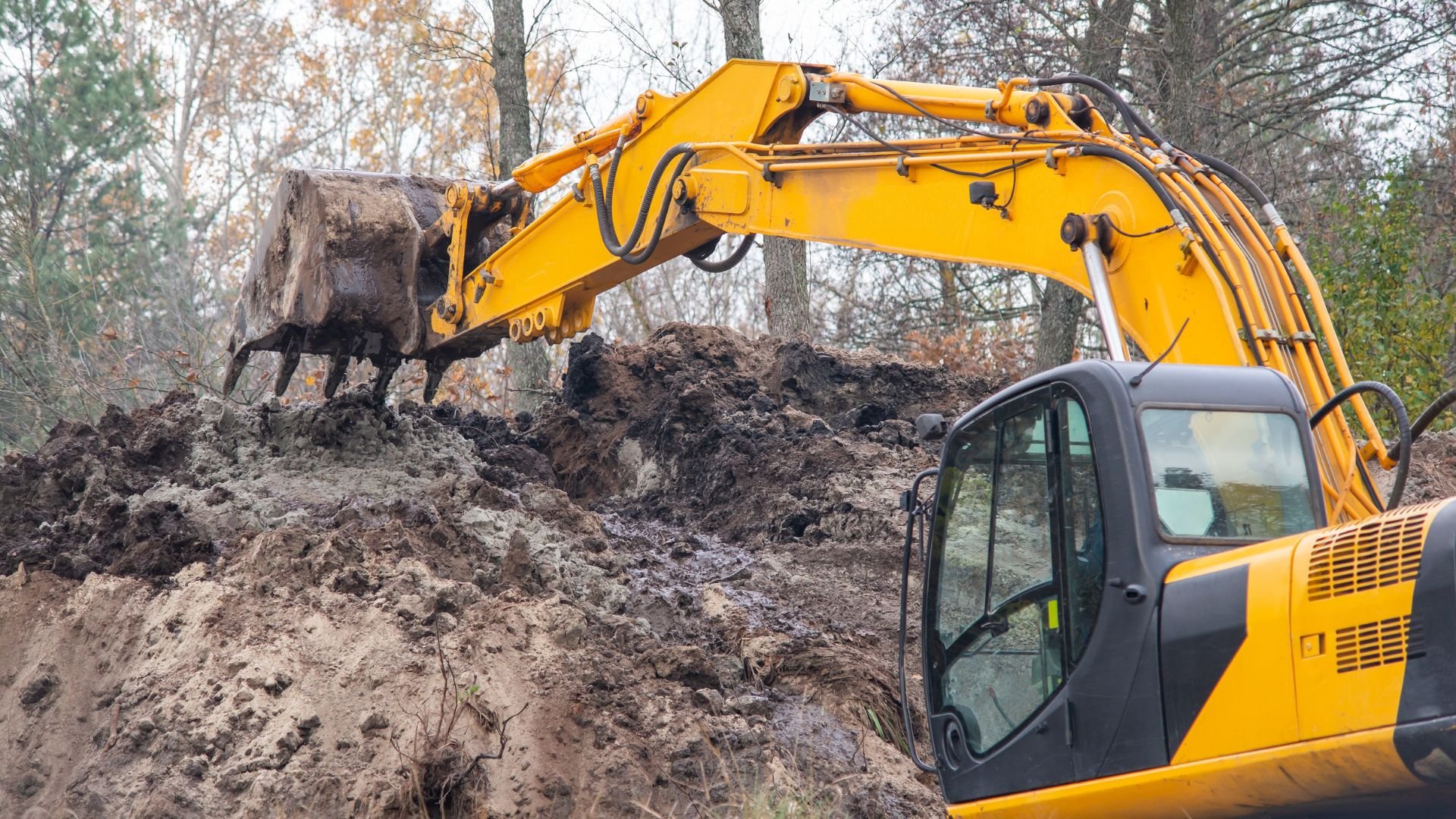 Yellow excavator digging large hole in muddy forest construction site