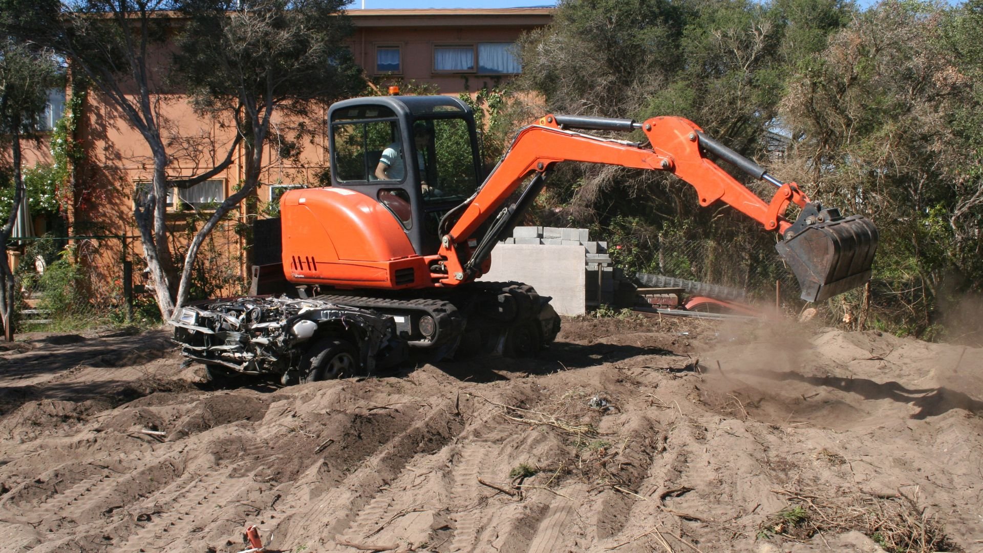 Orange excavator digging in dirt near residential building and trees